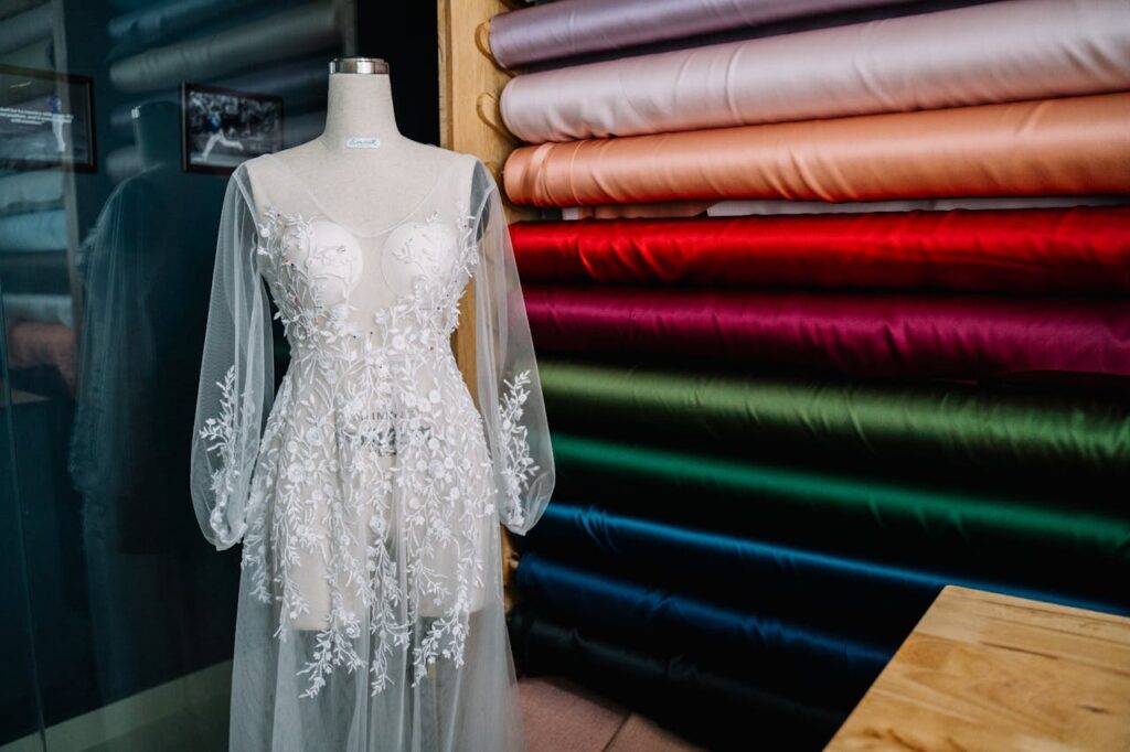 Intricately embroidered white wedding dress on a mannequin surrounded by colorful fabric bolts indoors.